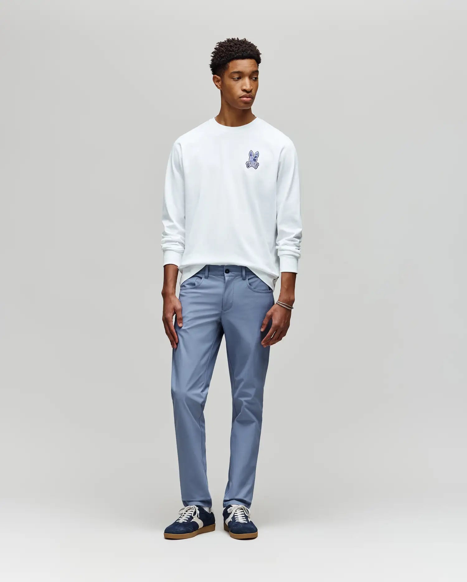 A young man stands against a plain light gray background, wearing the Psycho Bunny MENS ROWEN GRAPHIC TEE (MT0200103) in white, paired with light blue pants and black sneakers with white soles, looking slightly to his left.