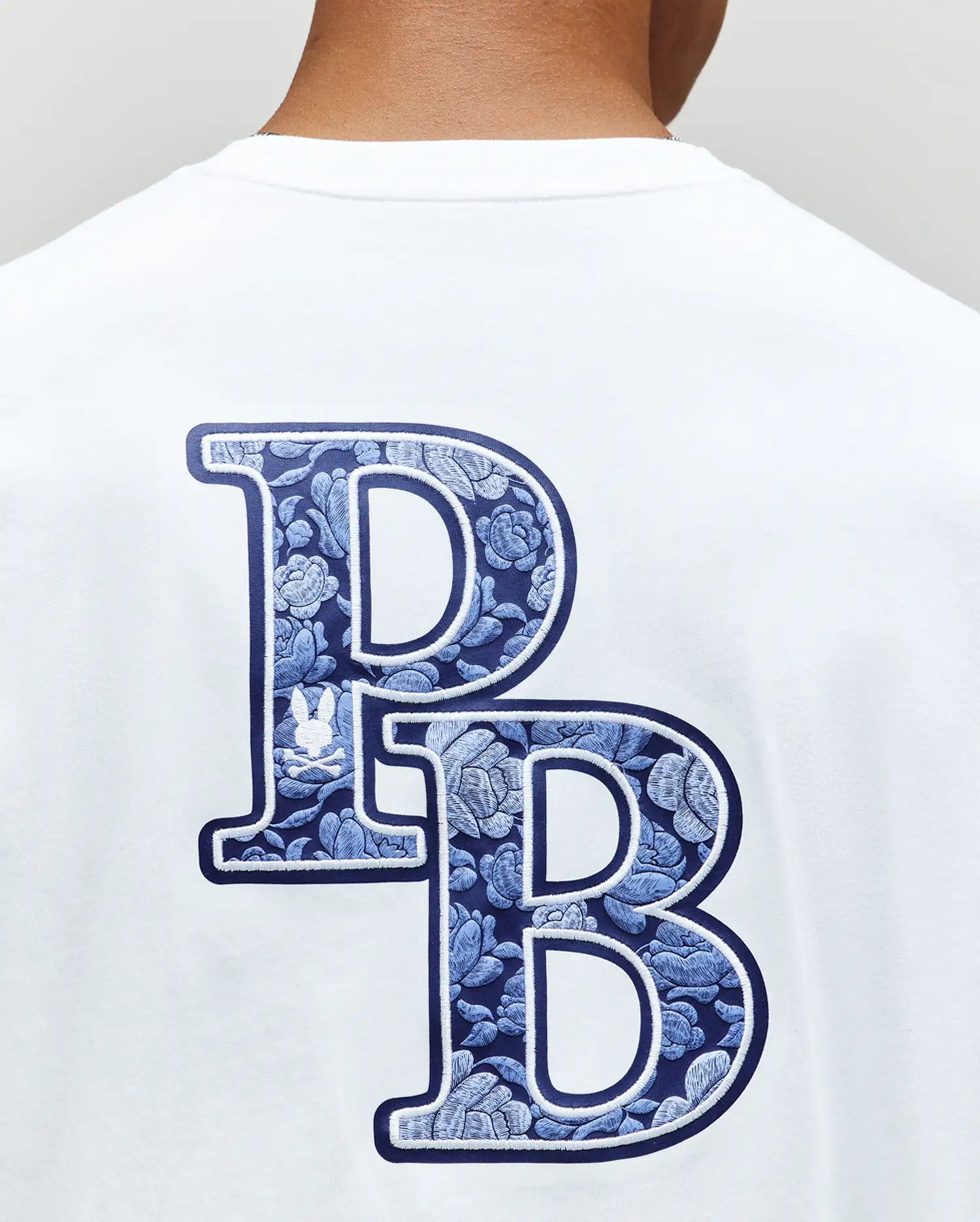 The Psycho Bunny MENS ROWEN GRAPHIC TEE (MT0200103) has large, overlapping blue "P" and "B" letters with a floral pattern on the back, plus a small white bunny logo inside the "P".