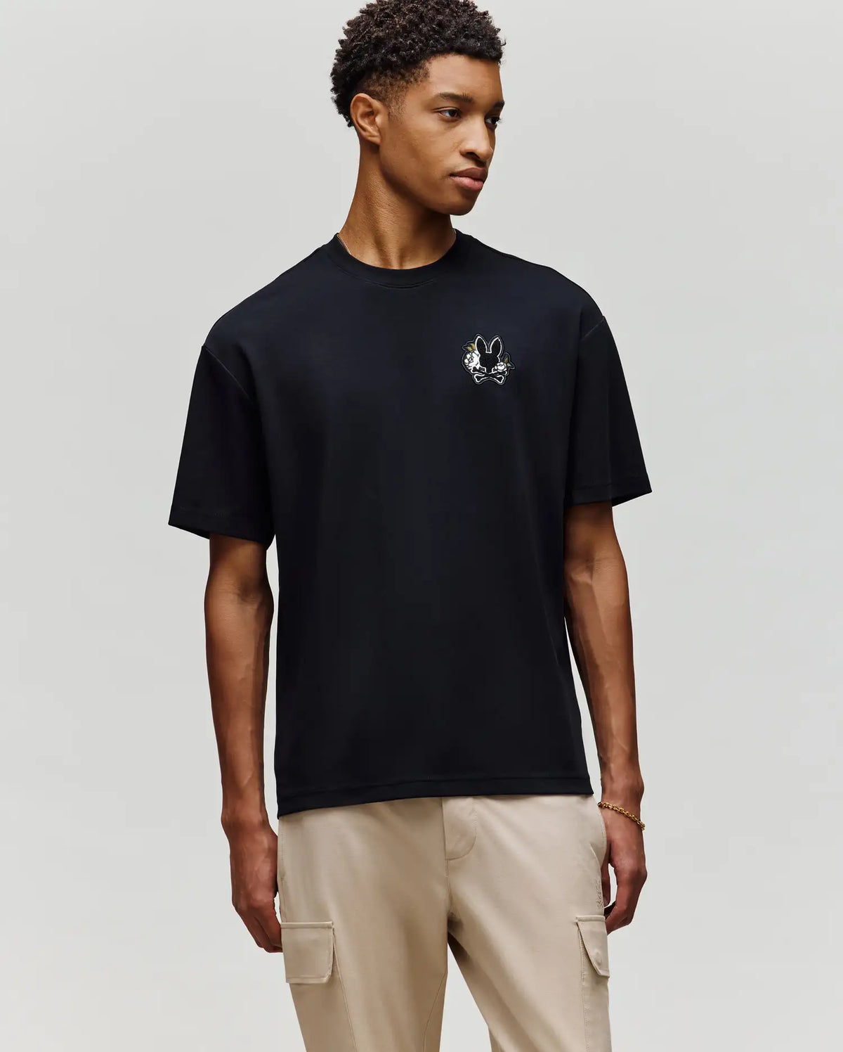 A young man wears the Psycho Bunny MENS AUGUSTE OVERSIZED TEE (MT0200104) featuring an embroidered Bunny logo, paired with light beige cargo pants for a laidback look against a plain gray background.