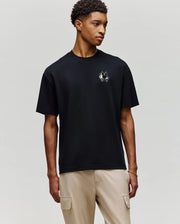 A young man wears the Psycho Bunny MENS AUGUSTE OVERSIZED TEE (MT0200104) featuring an embroidered Bunny logo, paired with light beige cargo pants for a laidback look against a plain gray background.