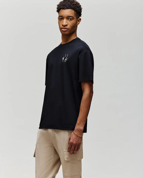 Against a plain light gray background, a person with short curly hair wears the Psycho Bunny MENS AUGUSTE OVERSIZED TEE (MT0200104) featuring an embroidered Bunny logo, paired with beige cargo pants for a relaxed, laidback look.