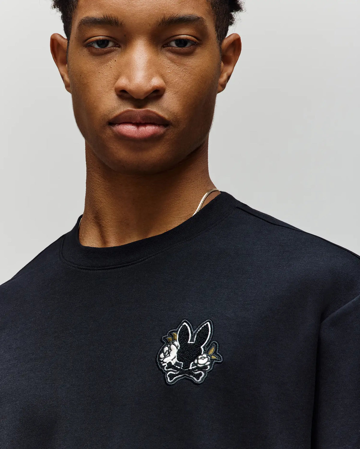 A person wearing the Psycho Bunny MENS AUGUSTE OVERSIZED TEE (MT0200104), featuring a small embroidered Bunny logo above two crossed carrots on the chest, ideal for a laidback style.