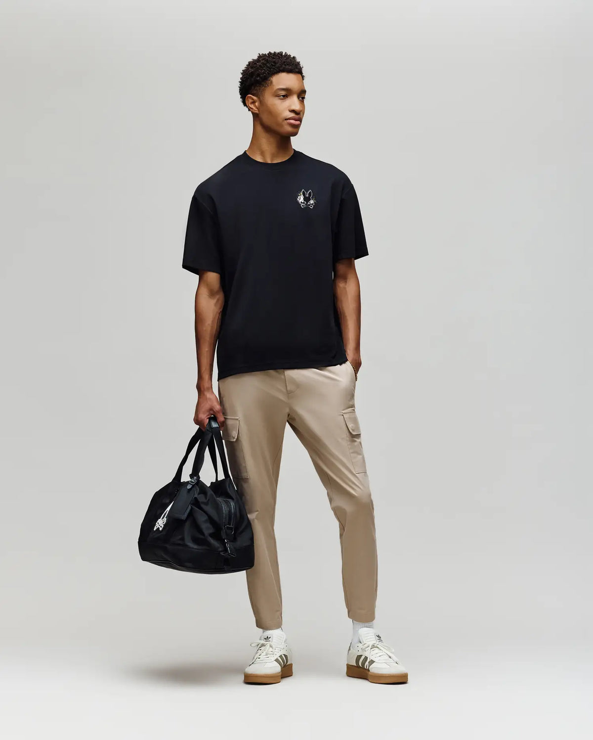 A young man stands against a plain background in the Psycho Bunny MENS AUGUSTE OVERSIZED TEE (MT0200104), paired with beige cargo pants, white sneakers, and holding a black duffel bag, embodying effortless laidback style.