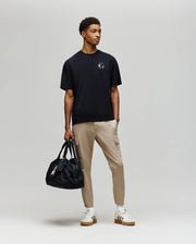 A young man stands against a plain background in the Psycho Bunny MENS AUGUSTE OVERSIZED TEE (MT0200104), paired with beige cargo pants, white sneakers, and holding a black duffel bag, embodying effortless laidback style.