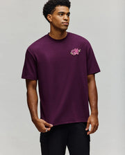 A man models the Psycho Bunny MENS AUGUSTE OVERSIZED TEE (MT0200104) in dark purple with a pink floral chest design, standing before a plain light gray background and highlighting its relaxed, laidback style.