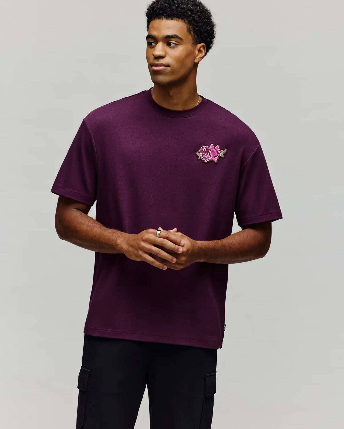 A man models the Psycho Bunny MENS AUGUSTE OVERSIZED TEE (MT0200104) in dark purple with a pink floral chest patch, standing against a light gray background and exuding effortless laidback style.