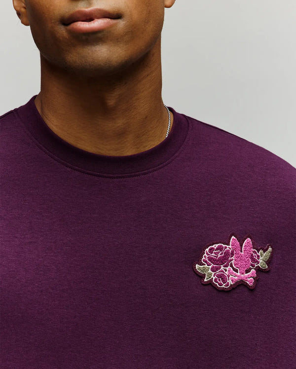 A person wears the Psycho Bunny MENS AUGUSTE OVERSIZED TEE (MT0200104) in dark purple, featuring a small pink and purple embroidered floral and bird patch on the left chest. Only the lower face and upper torso are shown, highlighting a laidback look.
