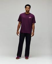 A man faces forward in a laidback style, wearing the Psycho Bunny MENS AUGUSTE OVERSIZED TEE (MT0200104) with a small embroidered Bunny logo, black cargo pants, and purple sneakers against a plain light gray background.