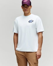 A man in the Psycho Bunny MENS AUGUSTE OVERSIZED TEE (MT0200104), tan cargo pants, and a purple floral cap stands against a gray background, exemplifying an effortless and laidback style.