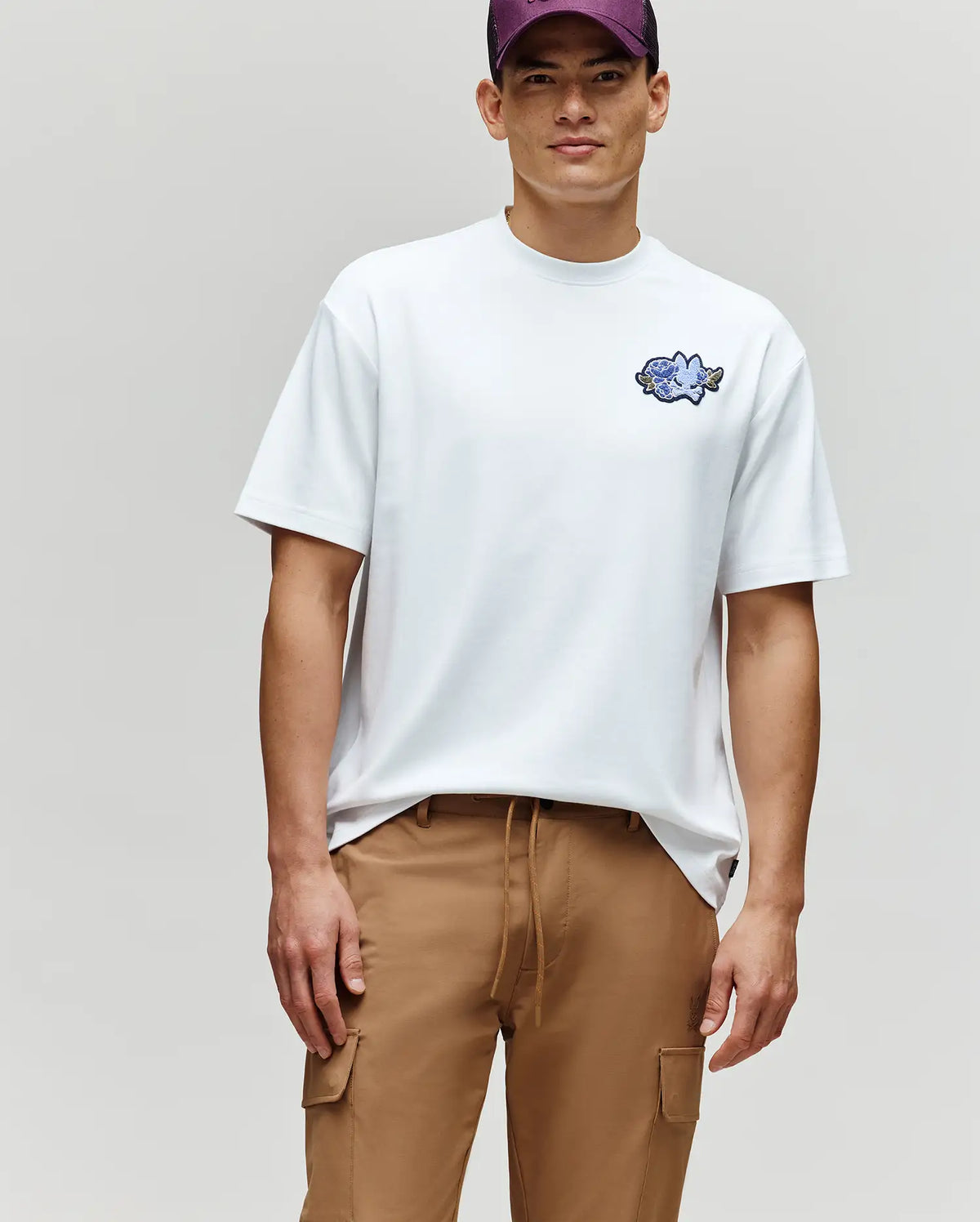 A man wears the Psycho Bunny MENS AUGUSTE OVERSIZED TEE (MT0200104) with a small embroidered Bunny logo, tan cargo pants, and a purple cap, standing against a plain light gray backdrop for a laidback look.