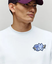 A man wears the Psycho Bunny MENS AUGUSTE OVERSIZED TEE (MT0200104) in white, featuring a blue floral chest patch and embroidered Bunny logo, styled with a purple baseball cap for a relaxed look.