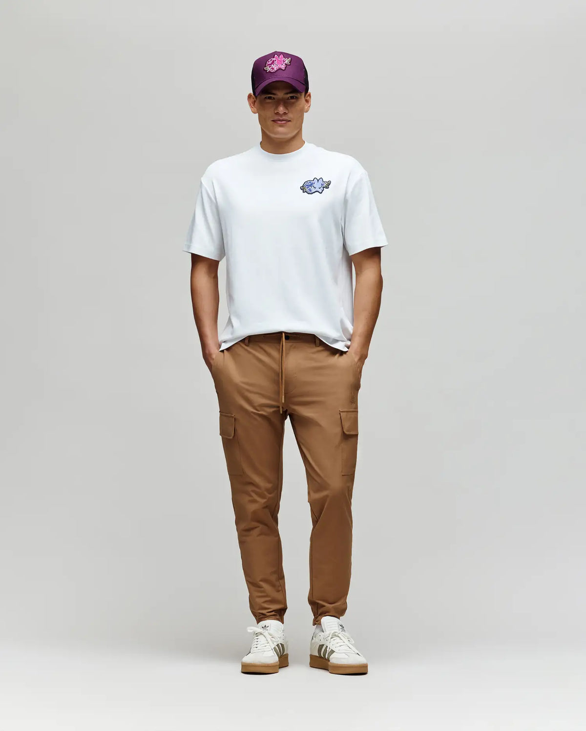 A person stands against a light gray background wearing the Psycho Bunny MENS AUGUSTE OVERSIZED TEE (MT0200104), tan cargo pants, white sneakers, and a purple graphic baseball cap—hands in pockets, looking at the camera.