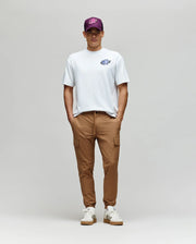 A person stands against a light gray background wearing the Psycho Bunny MENS AUGUSTE OVERSIZED TEE (MT0200104), tan cargo pants, white sneakers, and a purple graphic baseball cap—hands in pockets, looking at the camera.