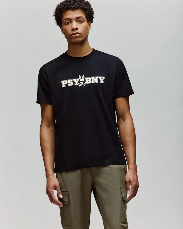A man stands against a plain background, wearing the Psycho Bunny MENS WADE GRAPHIC TEE - MT0200105 in black with "PSYBNY" and a bunny graphic on the chest, paired with olive green cargo pants.