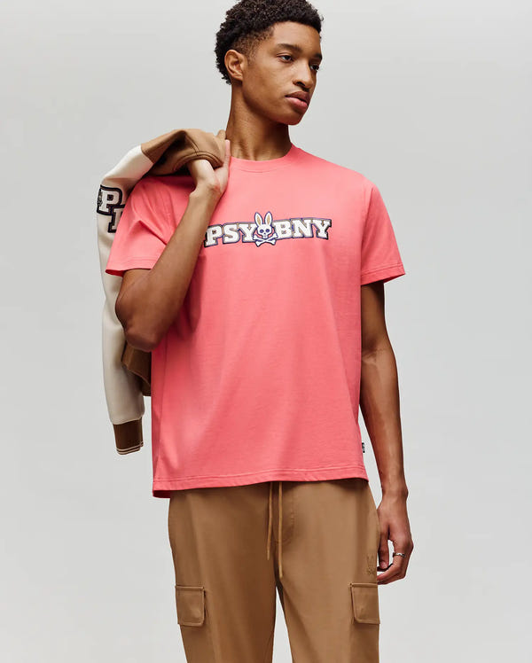 A young man wears the Psycho Bunny MENS WADE GRAPHIC TEE - MT0200105, a 100% cotton coral pink shirt with "PSYBUNNY" and a cartoon bunny graphic on the chest, tan cargo pants, and carries a beige and brown jacket over his shoulder.