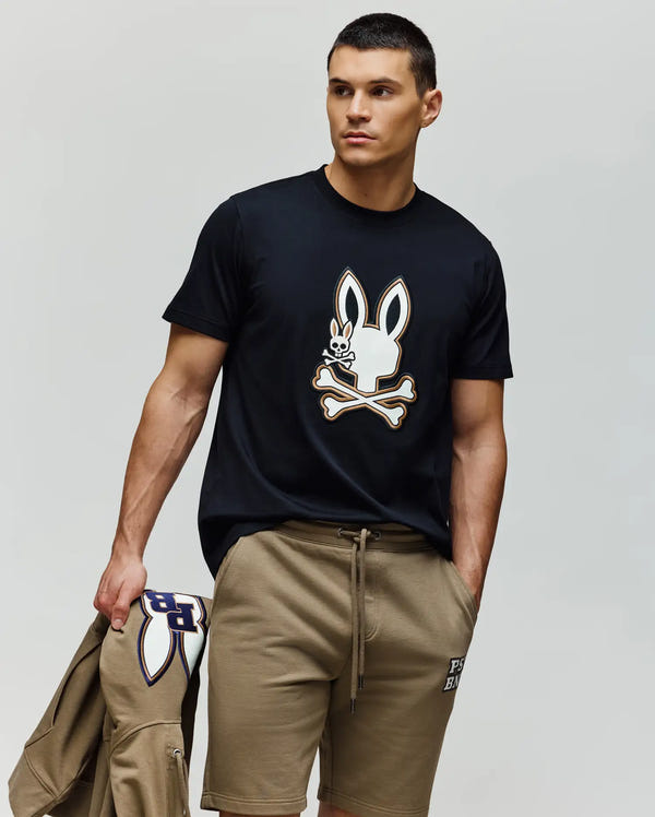 A man with short dark hair wears the Psycho Bunny MENS WADE GRAPHIC TEE - MT0200106, featuring an embroidered bunny and crossbones, paired with cotton khaki shorts. He carries a matching khaki jacket with the same motif, standing against a plain background.