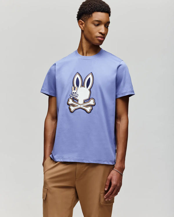 A young man wears the Psycho Bunny MENS WADE GRAPHIC TEE (MT0200106), a light purple 100% cotton shirt with an embroidered bunny and crossbones, paired with tan pants against a plain light background.