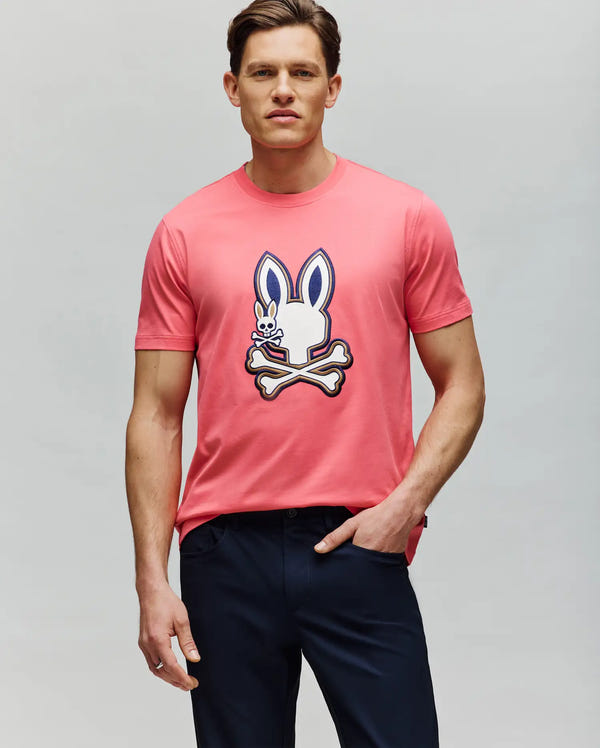 A man stands against a plain background wearing the Psycho Bunny MENS WADE GRAPHIC TEE - MT0200106, a pink 100% cotton t-shirt featuring a large embroidered Bunny head and crossbones graphic, paired with dark pants.