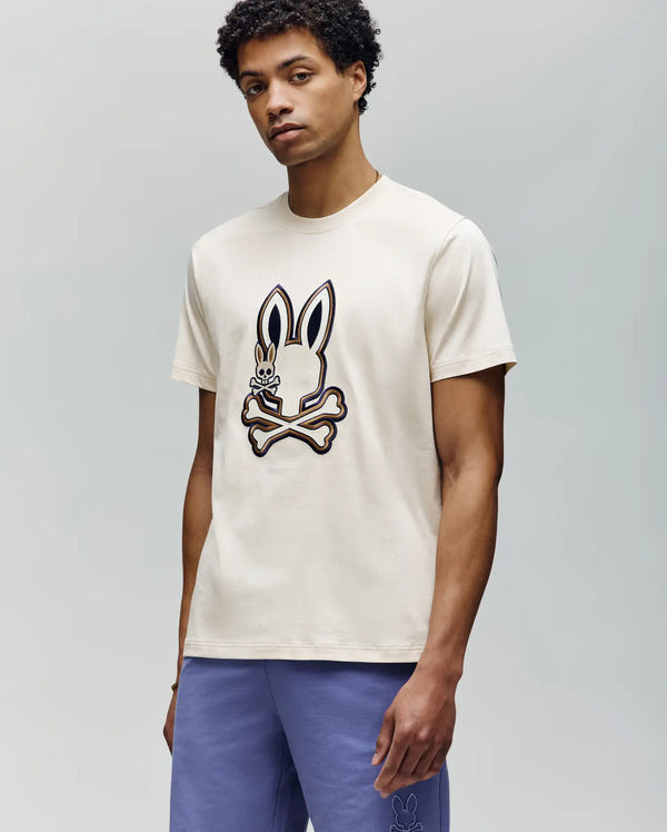 A man with short curly hair wears a cream-colored Psycho Bunny MENS WADE GRAPHIC TEE (MT0200106) featuring a large cartoon bunny with crossbones. He pairs it with light purple shorts, standing against a plain gray background.