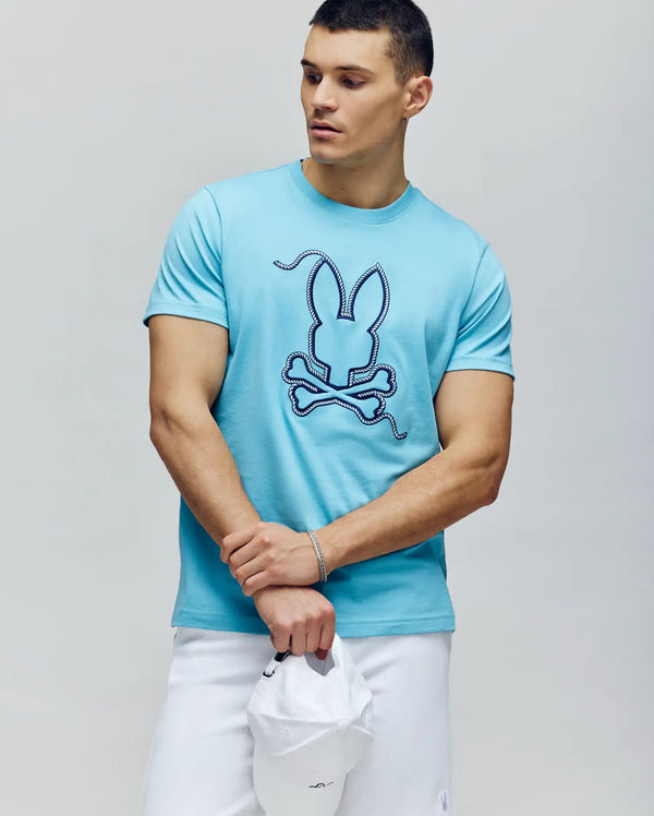 A man in the Psycho Bunny MENS CYRUS GRAPHIC TEE - MT0200115, featuring a stylized bunny and crossbones, stands with a white cap and shorts, looking left and embodying a relaxed sailor vibe.