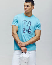 A man in the Psycho Bunny MENS CYRUS GRAPHIC TEE - MT0200115, featuring a stylized bunny and crossbones, stands with a white cap and shorts, looking left and embodying a relaxed sailor vibe.