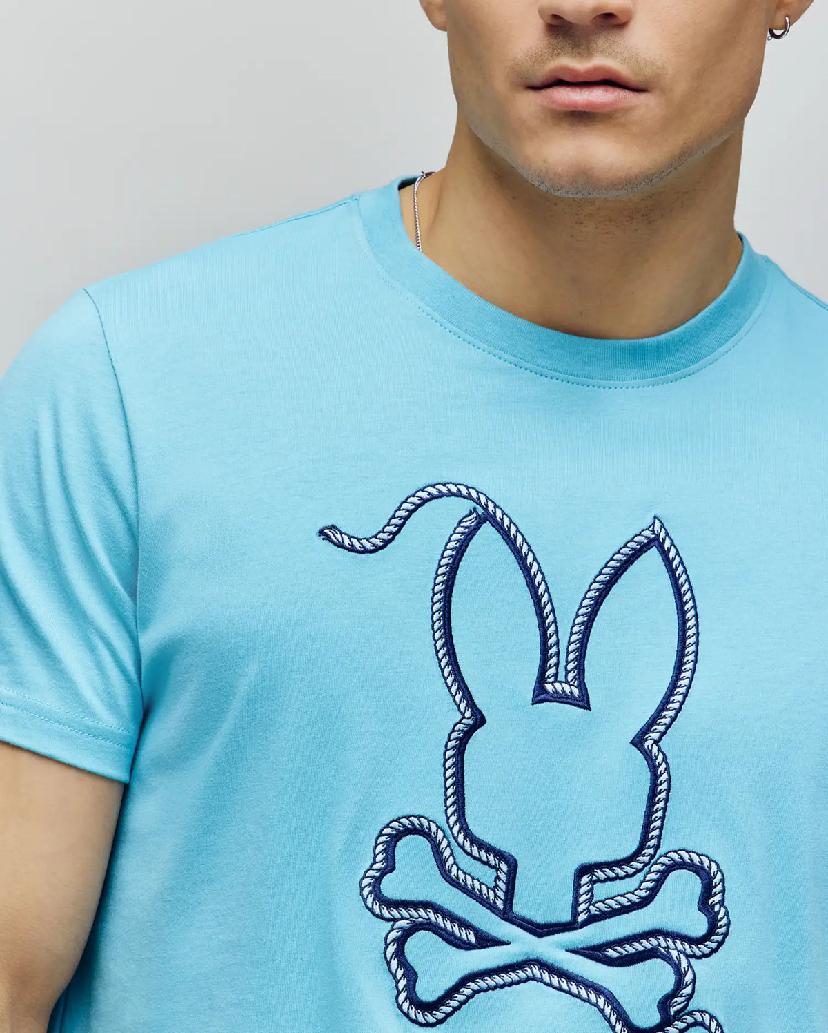 A man wears the Psycho Bunny MENS CYRUS GRAPHIC TEE (MT0200115) in light blue Pima cotton, featuring a navy rope-outlined bunny and crossbones. Only his lower face and upper torso are visible, evoking a sailor’s spirit.