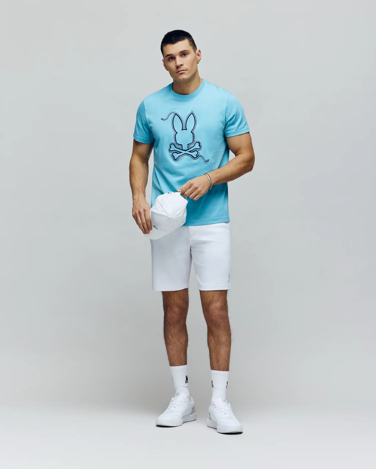 A man stands against a plain background wearing the Psycho Bunny MENS CYRUS GRAPHIC TEE - MT0200115 in light blue, paired with white shorts, white high-top sneakers, and holding a white baseball cap in his left hand.