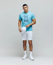 A man stands against a plain background wearing the Psycho Bunny MENS CYRUS GRAPHIC TEE - MT0200115 in light blue, paired with white shorts, white high-top sneakers, and holding a white baseball cap in his left hand.