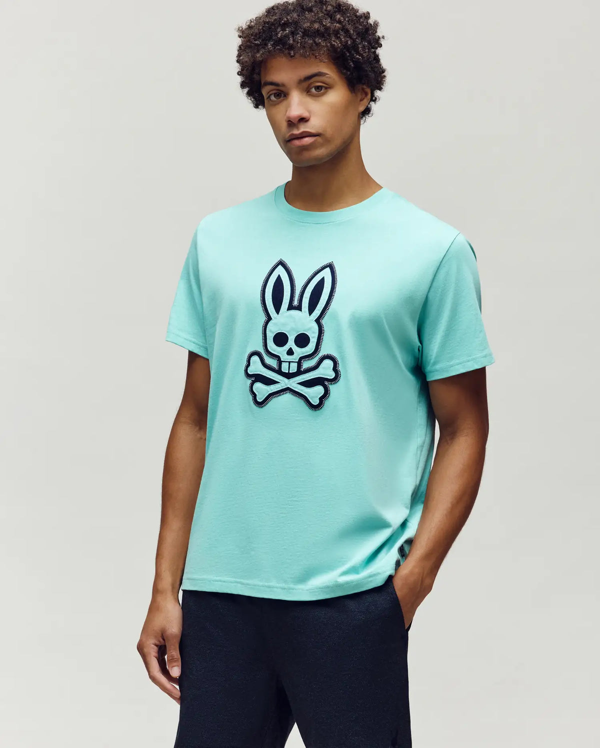 Mens Gage Graphic Tee, a light teal classic fit jersey t-shirt featuring a large embroidered navy Psycho Bunny skull and crossbones logo with white outline and zig-zag stitching on the chest.