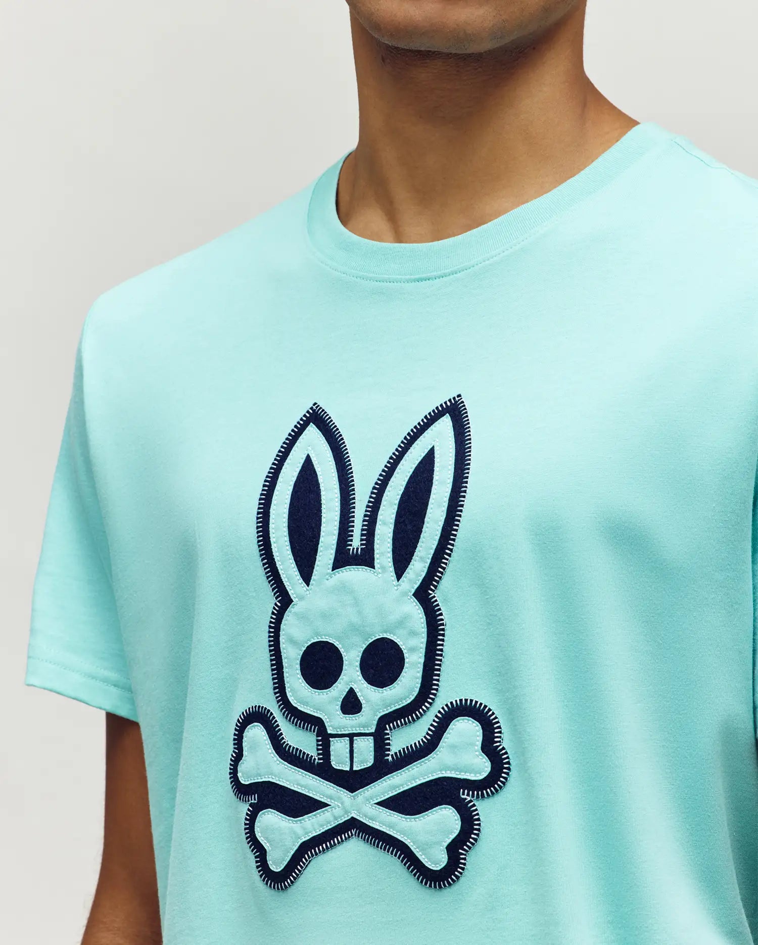 Mens Gage Graphic Tee. Aqua classic fit jersey t-shirt featuring a large navy blue and aqua appliqué Psycho Bunny skull and crossbones logo on the chest, detailed with white zigzag stitching.