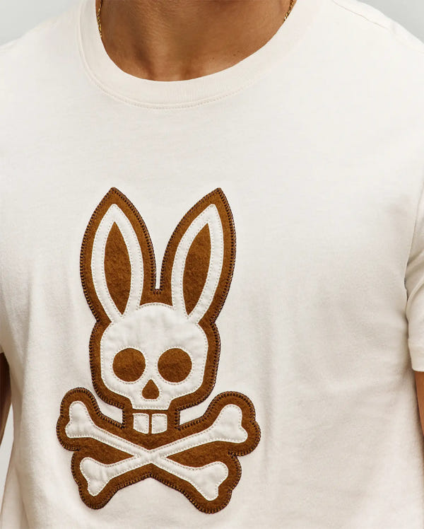 Mens Gage Graphic Tee, cream-colored jersey knit t-shirt featuring a prominent central brown felt and white stitched Psycho Bunny skull and crossbones appliqué, classic fit.