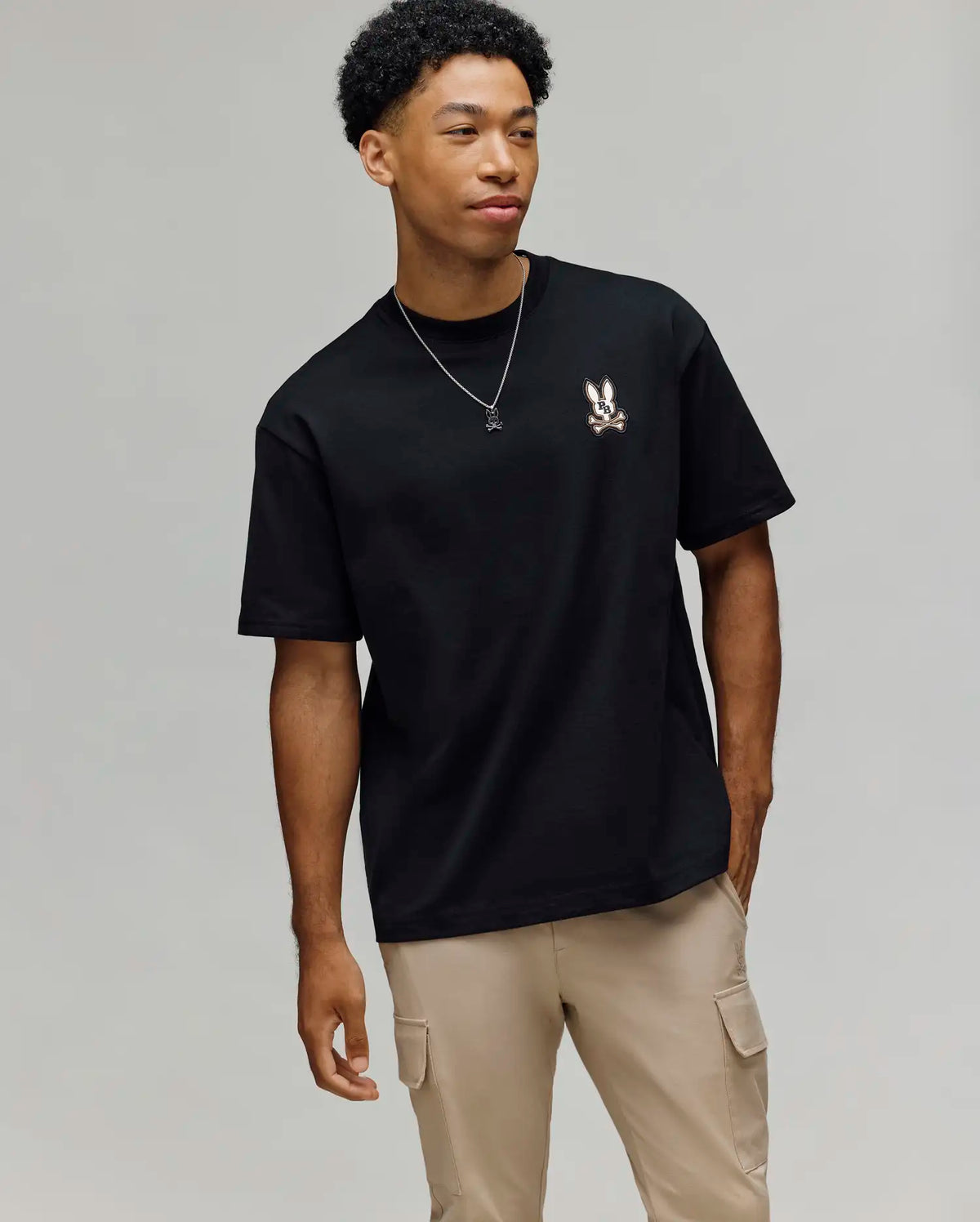A young man wears the Psycho Bunny MENS WADE OVERSIZED CHEST TEE (MT0200152), a relaxed fit black Pima cotton tee with a small bunny logo on the chest, paired with beige cargo pants, standing against a plain light background.