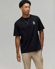 A young man wears the Psycho Bunny MENS WADE OVERSIZED CHEST TEE (MT0200152), a relaxed fit black Pima cotton tee with a small bunny logo on the chest, paired with beige cargo pants, standing against a plain light background.