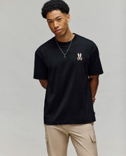 A young man with short curly hair wears the Psycho Bunny MENS WADE OVERSIZED CHEST TEE (MT0200152), a relaxed-fit black Peruvian Pima cotton shirt with a small bunny chest graphic, paired with beige pants and a necklace against a light grey background.