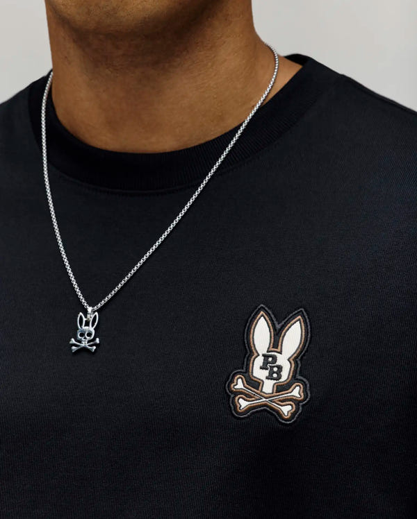 A person wearing the Psycho Bunny MENS WADE OVERSIZED CHEST TEE in black, featuring a relaxed fit and chest bunny and crossbones logo, paired with a silver chain necklace with a matching bunny and crossbones pendant.