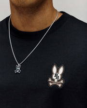 A person wearing the Psycho Bunny MENS WADE OVERSIZED CHEST TEE in black, featuring a relaxed fit and chest bunny and crossbones logo, paired with a silver chain necklace with a matching bunny and crossbones pendant.