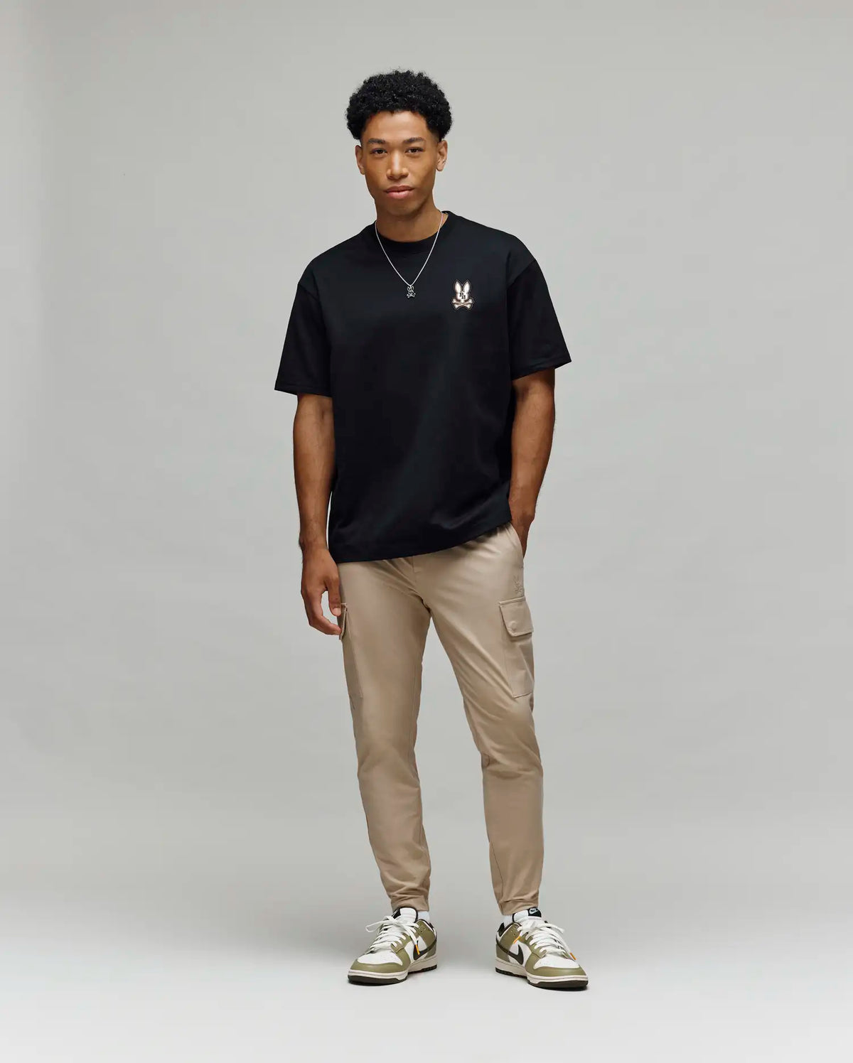 A man with short curly hair stands casually against a light gray background, wearing Psycho Bunny’s MENS WADE OVERSIZED CHEST TEE - MT0200152 in black, tan cargo pants, and white sneakers.