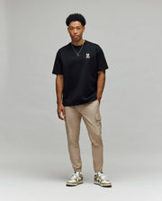 A man with short curly hair stands casually against a light gray background, wearing Psycho Bunny’s MENS WADE OVERSIZED CHEST TEE - MT0200152 in black, tan cargo pants, and white sneakers.