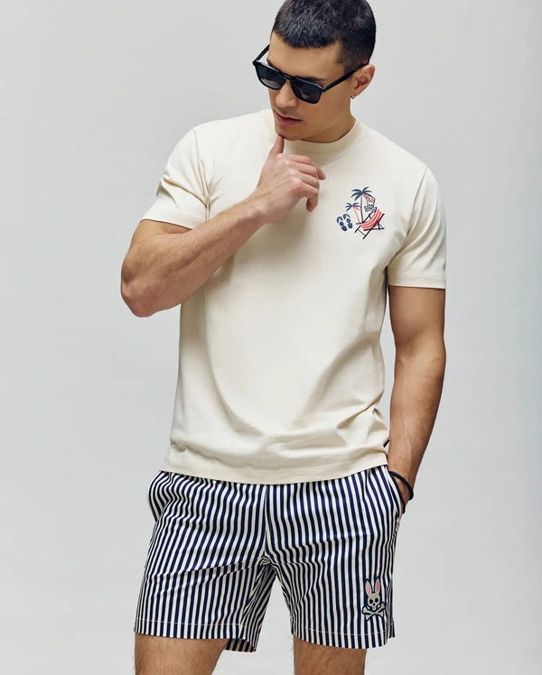 A man sports Psycho Bunny's MENS SONNY GRAPHIC TEE (MT0200174), a cream-colored Pima cotton shirt with a chest graphic, paired with striped shorts and sunglasses—ideal for a breathable summer look.