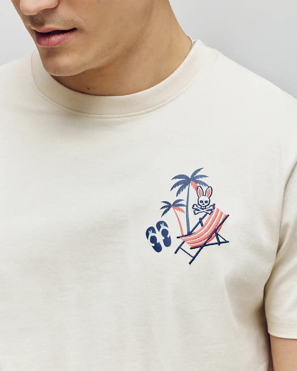 A person wearing the Psycho Bunny MENS SONNY GRAPHIC TEE (MT0200174), a cream Pima cotton shirt with a cartoon bunny lounging on a beach chair under palm trees—ideal for summer vibes.