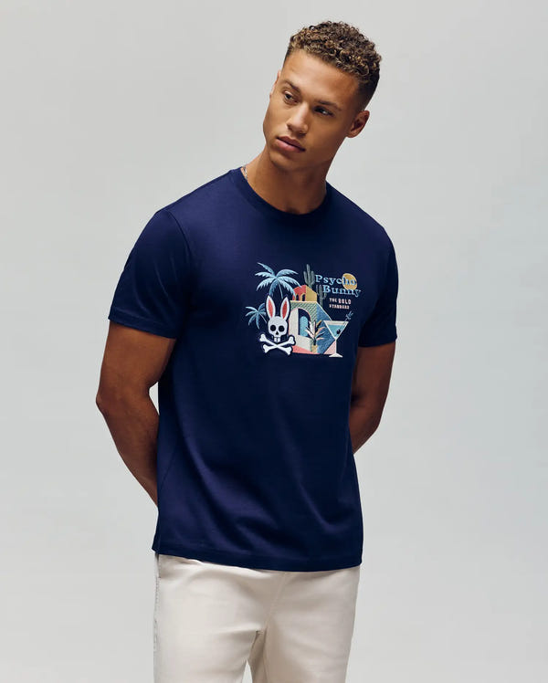 A young man with short curly hair wears the Psycho Bunny MENS SHANE GRAPHIC TEE - MT0200175, a navy blue summer tee with a cartoon rabbit, palm trees, colorful text, and martini glasses, paired with light pants against a gray background.