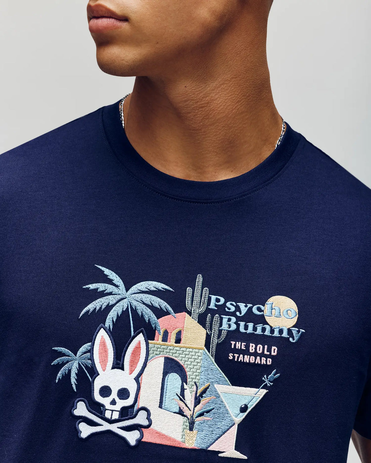 A man wears the Psycho Bunny MENS SHANE GRAPHIC TEE (MT0200175), a navy summer tee with colorful embroidery: palm trees, skull bunny logo, martini glasses, a cocktail, and "Psycho Bunny The Bold Standard" text.