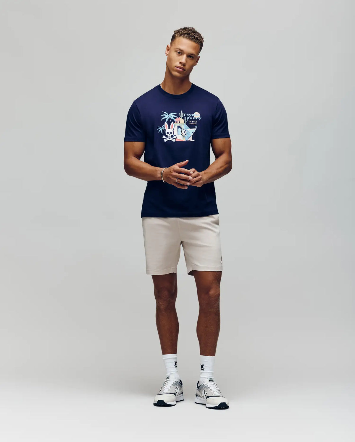 A man stands against a plain background wearing the Psycho Bunny MENS SHANE GRAPHIC TEE - MT0200175, featuring colorful martini glasses, paired with beige shorts, white crew socks, and white sneakers. He faces forward with hands clasped.