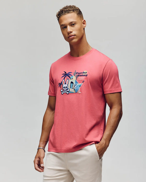 A man with short curly hair wears light-colored pants and the Psycho Bunny MENS SHANE GRAPHIC TEE (MT0200175), a pink Pima cotton summer tee with a cartoon graphic and text, standing before a plain light gray background.
