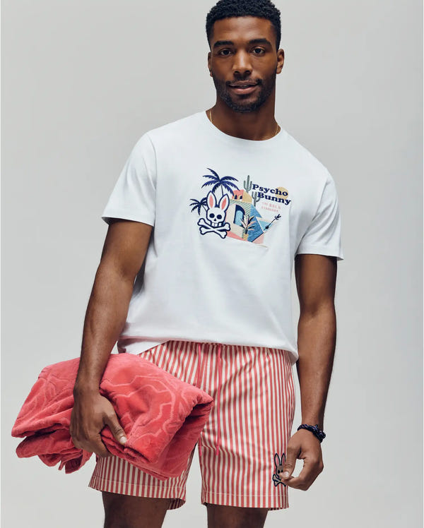 A man holds a folded red towel while wearing the Psycho Bunny MENS SHANE GRAPHIC TEE - MT0200175, a white Pima cotton summer tee with an embroidered graphic and text, paired with red-and-white striped shorts against a plain background.