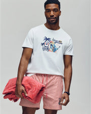 A man holds a folded red towel while wearing the Psycho Bunny MENS SHANE GRAPHIC TEE - MT0200175, a white Pima cotton summer tee with an embroidered graphic and text, paired with red-and-white striped shorts against a plain background.