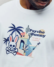 Close-up of the Psycho Bunny MENS SHANE GRAPHIC TEE, a soft Pima cotton summer tee with a vibrant embroidered skull-and-crossbones bunny logo, palm trees, cacti, geometric buildings, and "THE GOLD STANDARD" text.