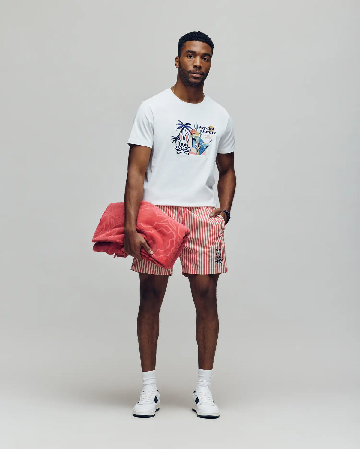 A man stands against a plain background wearing the Psycho Bunny MENS SHANE GRAPHIC TEE - MT0200175 in white, paired with red and white striped shorts, white sneakers, and holding a large red towel. He looks forward with a neutral expression.