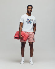 A man stands against a plain background wearing the Psycho Bunny MENS SHANE GRAPHIC TEE - MT0200175 in white, paired with red and white striped shorts, white sneakers, and holding a large red towel. He looks forward with a neutral expression.