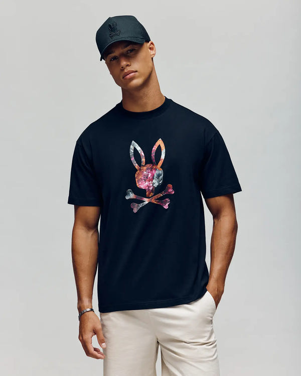 A young man wears the Psycho Bunny MENS NOVAN GRAPHIC TEE - MT0200177 in black, featuring a colorful floral skull and crossbones bunny graphic, paired with a black cap and light pants, standing against a plain light background.